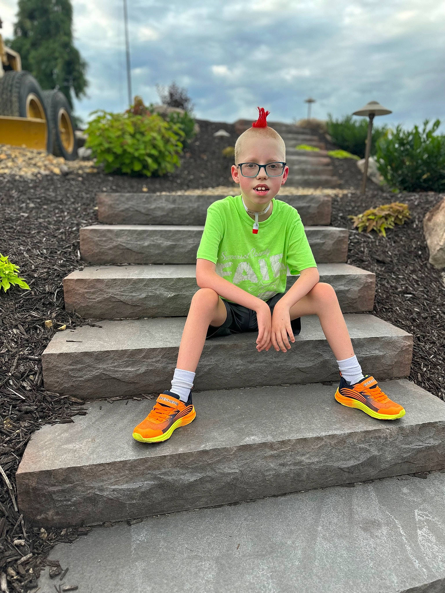 Carson is registered to the contest to win money with this photo: asphalt, cloud, eyewear, fun, grass, human_leg, knee, leisure, person, plant, recreation, road_surface, shorts, sidewalk, sky, sneakers, sportswear, t_shirt, thigh, toddler