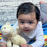 Angad is registered to the contest to win money with this photo: baby, blanket, bracelet, child, curly_hair, cute, expression, face, floor, hand, indoor, infant, looking_at_camera, lying_down, portrait, small_child, soft_toy, teddy_bear, toy, white_clothing