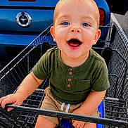 Colton is registered to the contest to win money with this photo: baby, child, toddler, smiling, happy, portrait, blue_eyes, outdoor, shopping_cart, grocery_cart, car, blue_car, parking_lot, sitting, green_shirt, shorts, infant, face, metal, daylight