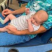Ryker is registered to the contest to win money with this photo: baby, beard, blue_shirt, camping_chair, casual, chair, daylight, face, family, greenery, hand, holding, man, outdoor, parenting, relaxing, seated, socks, striped_onesie, wooden_floor
