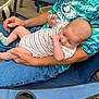 baby, beard, blue_shirt, camping_chair, casual, chair, daylight, face, family, greenery, hand, holding, man, outdoor, parenting, relaxing, seated, socks, striped_onesie, wooden_floor