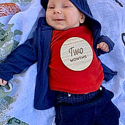 Ryker is registered to the contest to win money with this photo: baby, infant, smiling, clothing, jacket, pants, red_shirt, wooden_plaque, two_months, blanket, patterned_blanket, lying_down, happy, cute, face, hand, headwear, cozy, child, portrait