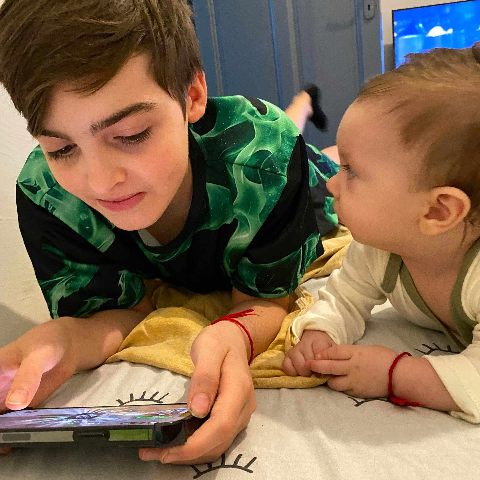 Jovano Et Angélo participe au concours pour gagner de l'argent avec cette photo : child, baby, bed, blanket, smartphone, red_bracelet, clothing, face, hand, person, indoor, technology, looking, lying_down, curly_hair, short_hair, casual, focus, playful, interaction