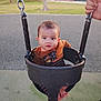 baby, child, swing, playground, park, outdoor, grass, tree, fence, person, hand, foot, clothing, chain, seat, expression, face, curious, daylight, recreation