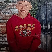 Julian is registered to the contest to win money with this photo: child, boy, smile, red_hoodie, sportswear, san_francisco_49ers, brick_wall, wooden_fence, outdoor, portrait, casual_clothing, happy, young_person, seated, hands_clasped, short_hair, daylight, background, expression, clothing