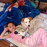 child, dog, pajamas, blanket, mattress, indoor, relaxed, smiling, sleeping, person, wood_floor, furniture, blue_blanket, pink_clothing, animal, pet, cute, happy, tongue_out, home