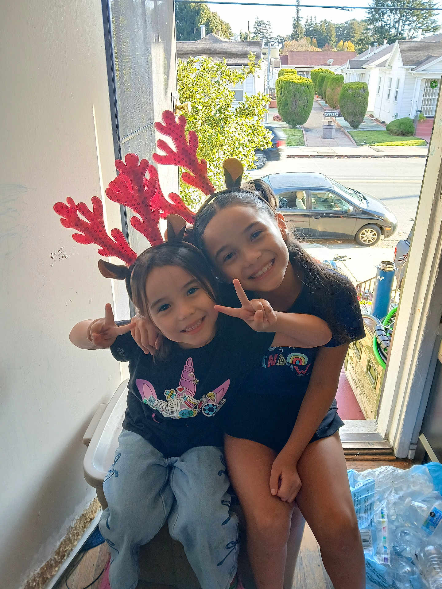 Vanessa is registered to the contest to win money with this photo: child, children, smiling, peace_sign, reindeer_antlers, headband, indoor, doorway, bench, casual_clothing, happy, sitting, portrait, two_people, daylight, window, street, car, trees, neighborhood