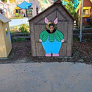 Vanessa is registered to the contest to win money with this photo: child, playhouse, face_cutout, outdoor, park, playground, cartoon_painting, bunny_ears, casual_clothing, green_shirt, blue_overalls, wooden_structure, plants, trees, sunny, daytime, fun, curious, amusement_park, colorful