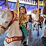 amusement_park, carousel, casual_clothing, child, daytime, decorated_horse, family, festival, fun, green_pants, happy, horse, leisure, outdoor, people, playful, purple_canopy, ride, smiling, yellow_top