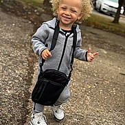 Dakarai is registered to the contest to win money with this photo: blonde_hair, casual_wear, child, crossbody_bag, curly_hair, daylight, fashion, happy, nike, outdoor, pavement, person, smiling, standing, street, toddler, tracksuit, urban, white_sneakers, young_child