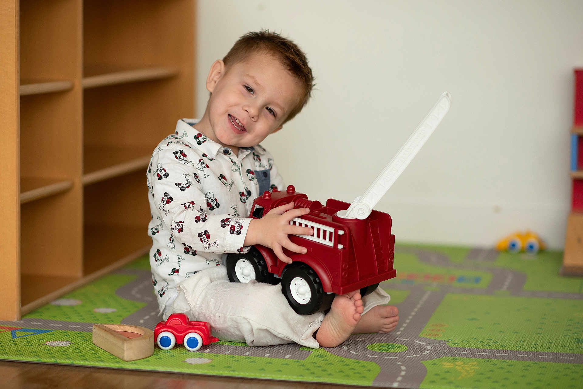 Timéo participe au concours pour gagner de l'argent avec cette photo : toddler, child, toy, fire_truck, play_mat, smiling, indoor, casual_clothing, wooden_shelves, barefoot, floor, playtime, cute, happy, young_child, room, colorful, head_tilt, small_toy_car, white_pants