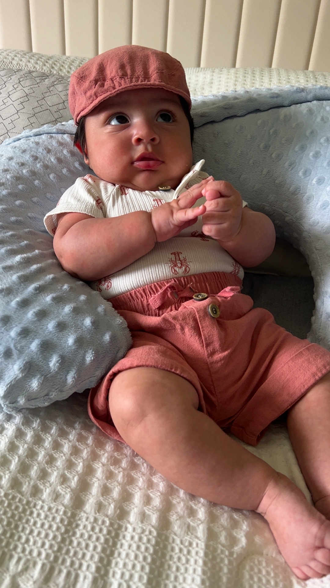 Noah is registered to the contest to win money with this photo: baby, infant, cap, pink_clothing, shorts, pillow, bed, blanket, texture, hands, face, curious, cute, child, indoors, soft_lighting, skin, portrait, sitting, cozy