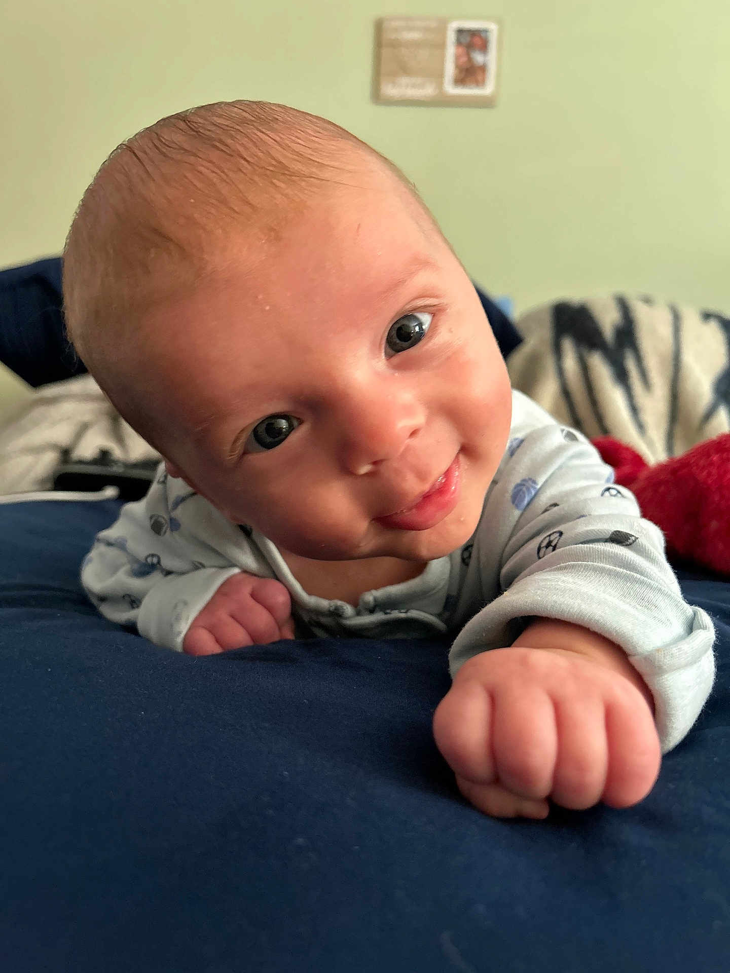 Asher is registered to the contest to win money with this photo: baby, infant, smiling, face, hand, fist, blue_clothing, lying_down, indoors, bed, blanket, head, eyes, skin, cute, portrait, young_child, happy, closeup, person