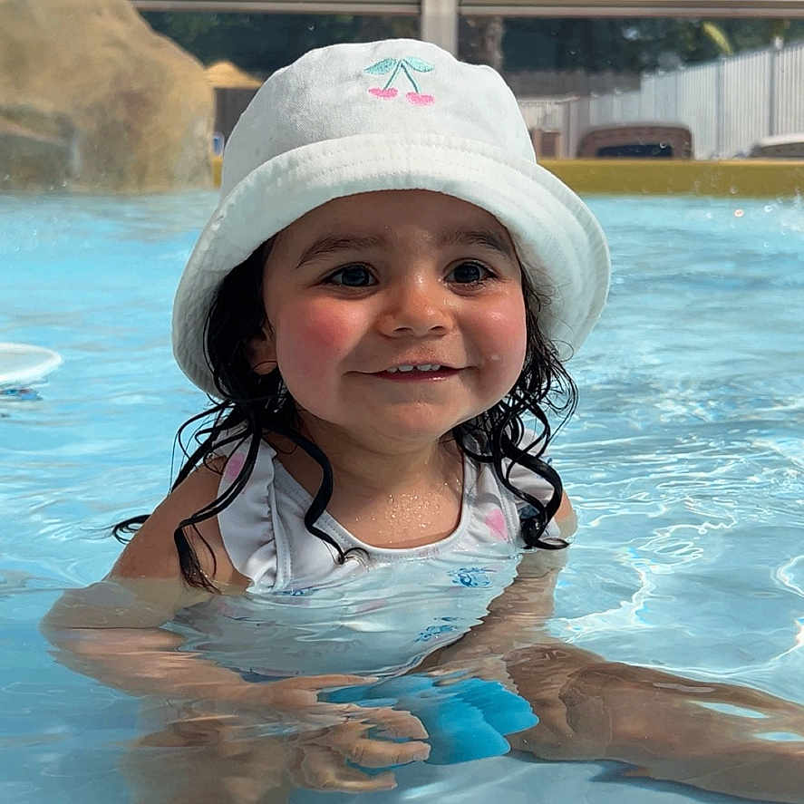 Mia a rejoint le concours — aidez-le/la à gagner de superbes lots ! blue_sky, bucket_hat, child, clothing, cute, face, happy, hat, outdoor, palm_tree, person, pool, smiling, splashing, summer, sunny, swimming, toddler, toy, water
