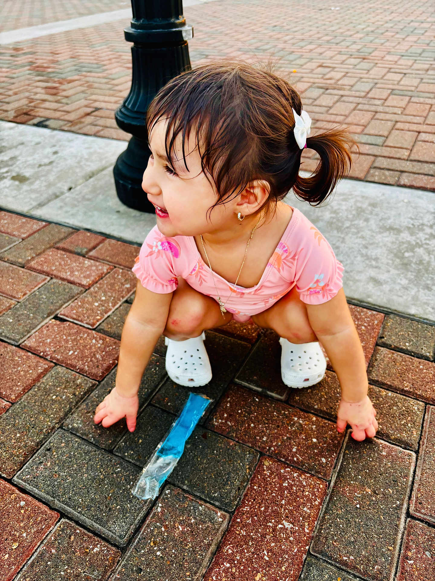 Mailyna joined the competition — help win amazing prizes! toddler, child, girl, wet_hair, ponytail, pink_clothing, white_bow, white_crocs, jewelry, bracelet, pavement, brick_floor, lamp_post, outdoor, smiling, crouching, playful, summer, popsicle_wrapper, happy