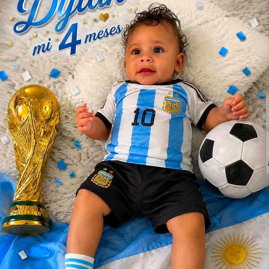 Dylan joined the competition — help win amazing prizes! baby, child, soccer_ball, trophy, confetti, blanket, blue, white, football_jersey, socks, shorts, cute, portrait, celebration, sports, argentina_flag, indoor, smiling, young_child, lying_down