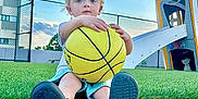 Jack is registered to the contest to win money with this photo: child, basketball, grass, playground, slide, outdoor, blue_sky, clouds, blonde_hair, blue_eyes, sitting, shoes, concrete, fence, building, summer, daylight, casual_clothing, young_child, recreation