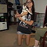 woman, person, cat, pet, animal, indoors, kneeling, smiling, glasses, casual_clothing, shorts, t_shirt, hugging, pet_carrier, cage, living_room, furniture, slipper, leopard_print, portrait