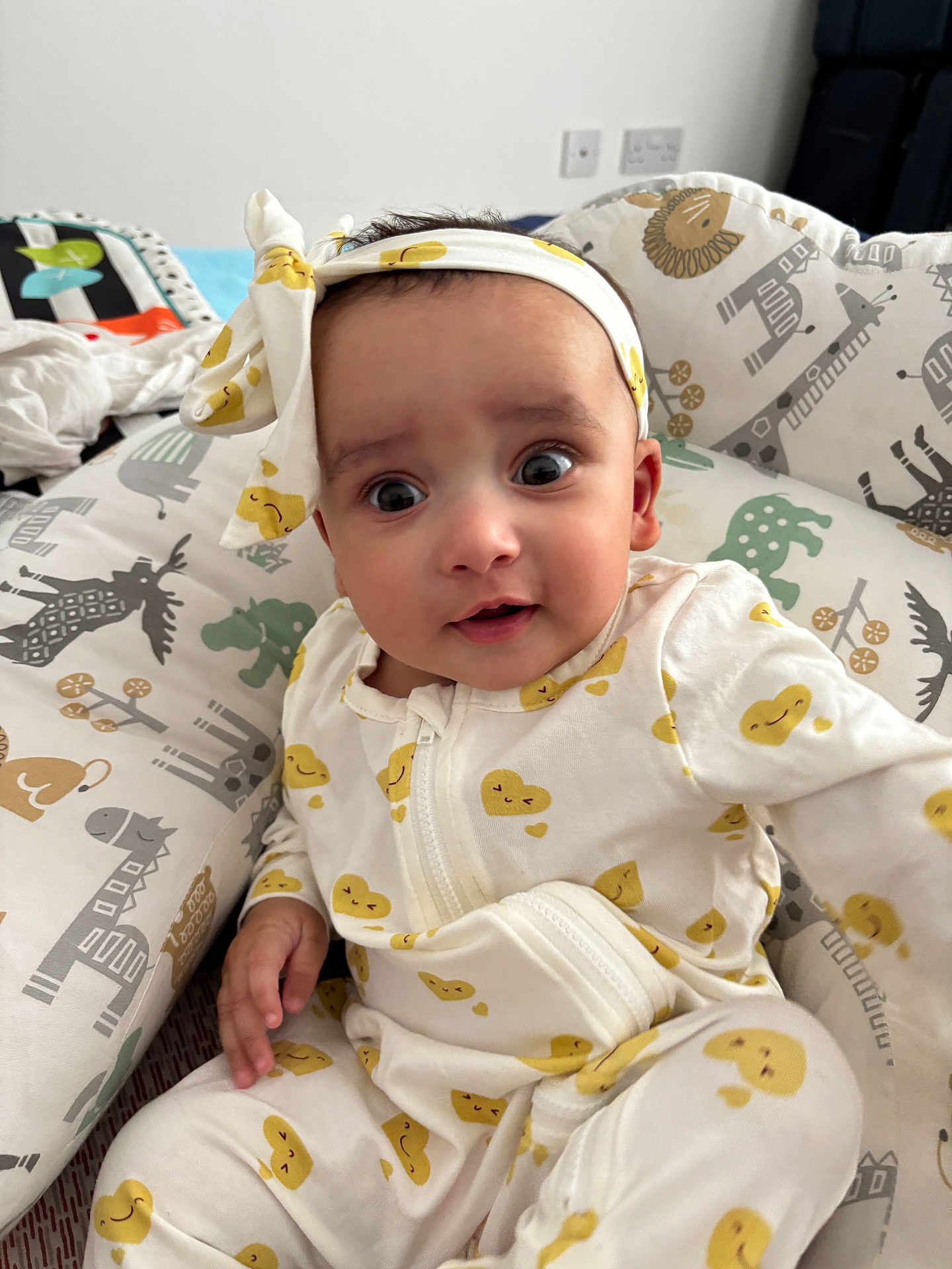Praavi is registered to the contest to win money with this photo: baby, infant, child, headband, yellow_hearts, onesie, cushion, animal_print, giraffe, elephant, zebra, bed, bedding, curious_expression, indoors, portrait, cute, soft, cozy, smiling