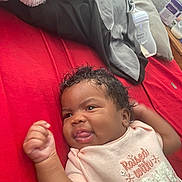 Ryann joined the competition — help win amazing prizes! baby, infant, curly_hair, pink_clothing, bed, red_bedspread, blanket, baby_bottle, smiling, happy, lying_down, fist_raised, cozy, indoors, cute, child, portrait, soft_lighting, expression, young