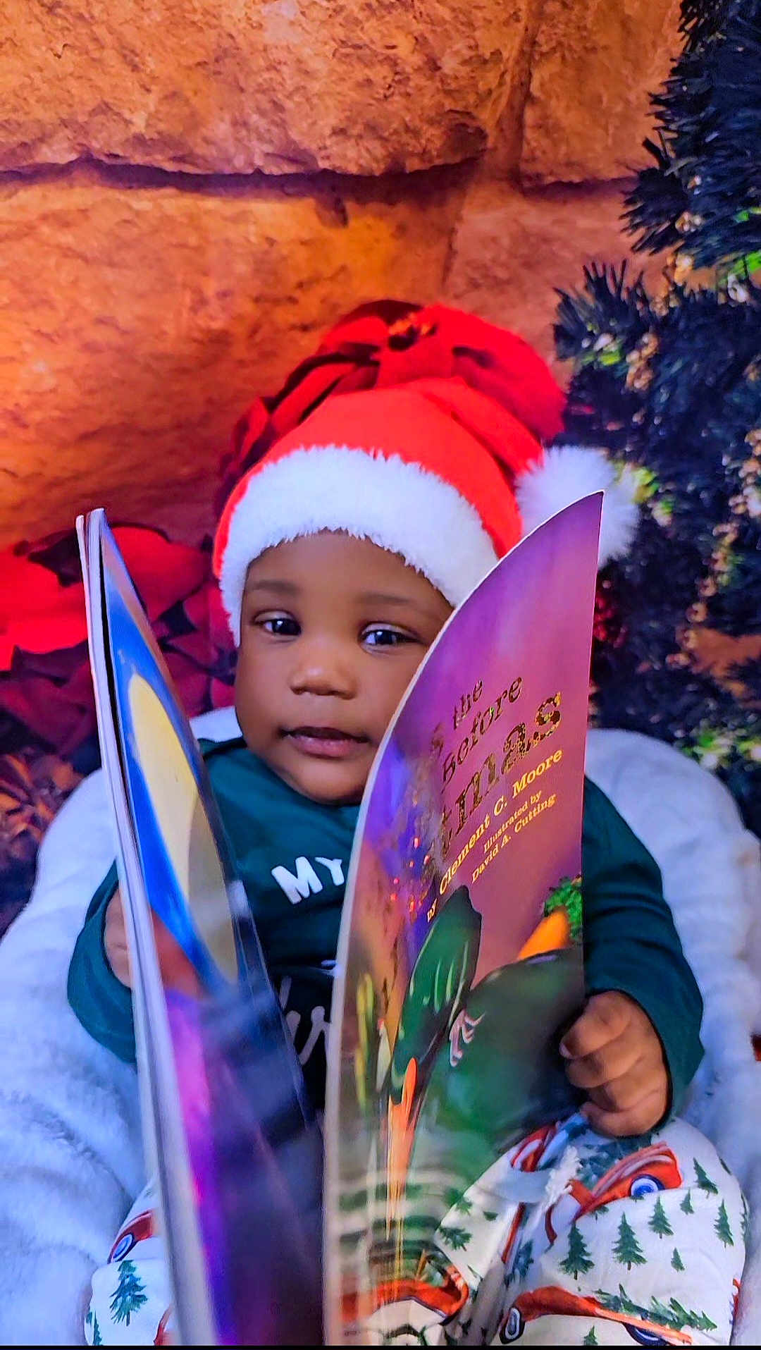 Noah is registered to the contest to win money with this photo: toddler, child, santa_hat, book, reading, holiday, christmas, festive, cozy, indoor, pajamas, smiling, face, person, decorations, tree, winter, cute, happy, warm