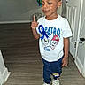 child, boy, toddler, portrait, standing, thumbs_up, smiling, indoors, hallway, door, balloons, birthday, t_shirt, cartoon, sneakers, blue_pants, wood_floor, home, celebration, face