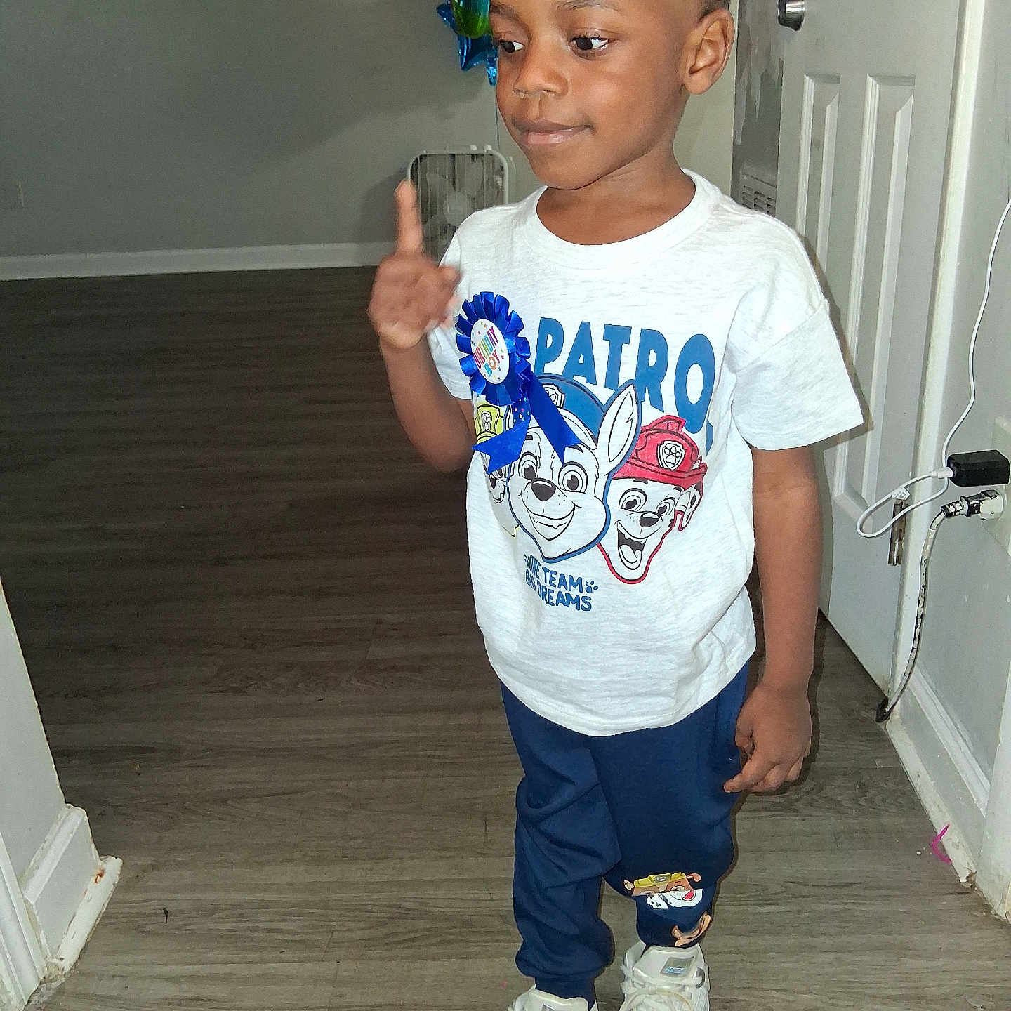 Jayceon is registered to the contest to win money with this photo: child, boy, toddler, portrait, standing, thumbs_up, smiling, indoors, hallway, door, balloons, birthday, t_shirt, cartoon, sneakers, blue_pants, wood_floor, home, celebration, face