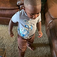Aiden is registered to the contest to win money with this photo: toddler, child, standing, indoors, living_room, leather_chair, carpet, toy, astronaut_shirt, brown_pants, short_hair, looking_down, walking, portrait, home, furniture, floor, casual_clothing, cute, daylight