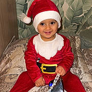 Kaïs a rejoint le concours — aidez-le/la à gagner de superbes lots ! toddler, child, santa_claus_costume, red_clothing, hat, smiling, indoor, bedspread, patterned_fabric, phone, beaded_chain, holiday, festive, cute, happy, portrait, person, wearing_hat, sitting, home