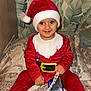 toddler, child, santa_claus_costume, red_clothing, hat, smiling, indoor, bedspread, patterned_fabric, phone, beaded_chain, holiday, festive, cute, happy, portrait, person, wearing_hat, sitting, home