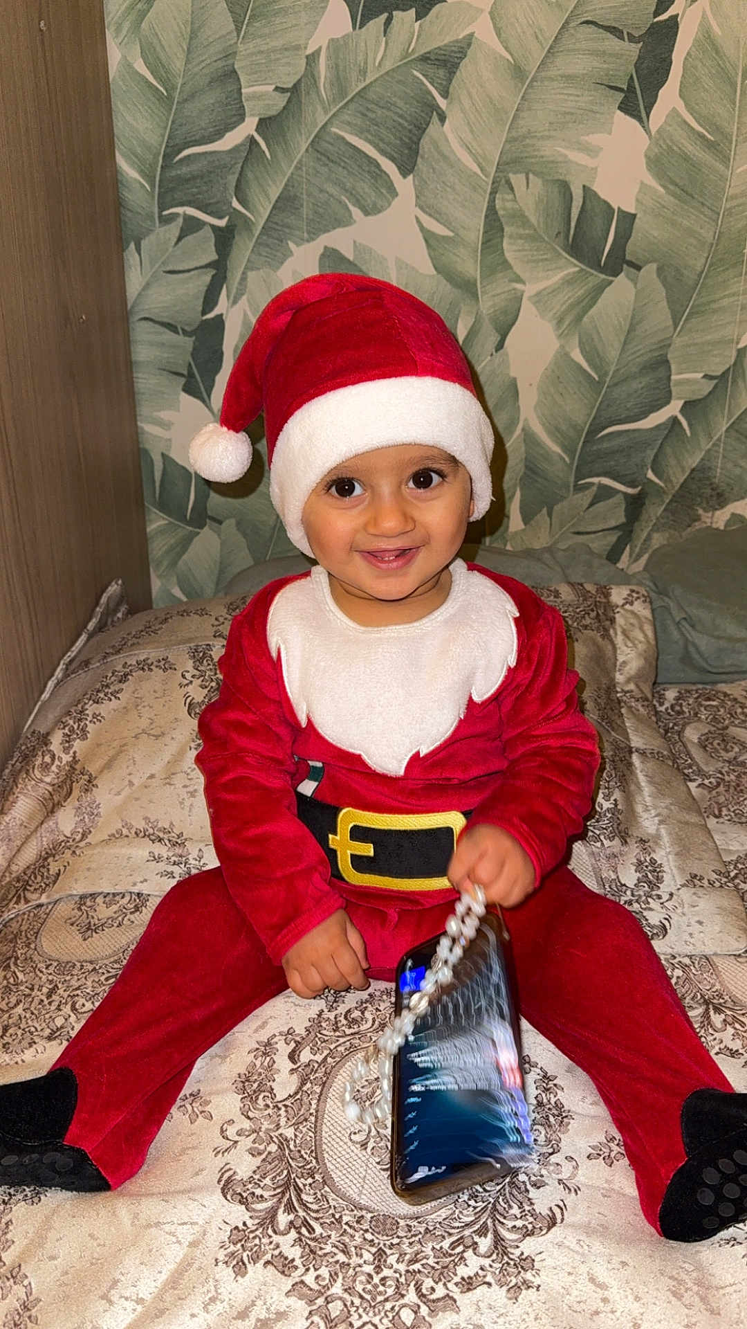 Kaïs a rejoint le concours — aidez-le/la à gagner de superbes lots ! toddler, child, santa_claus_costume, red_clothing, hat, smiling, indoor, bedspread, patterned_fabric, phone, beaded_chain, holiday, festive, cute, happy, portrait, person, wearing_hat, sitting, home