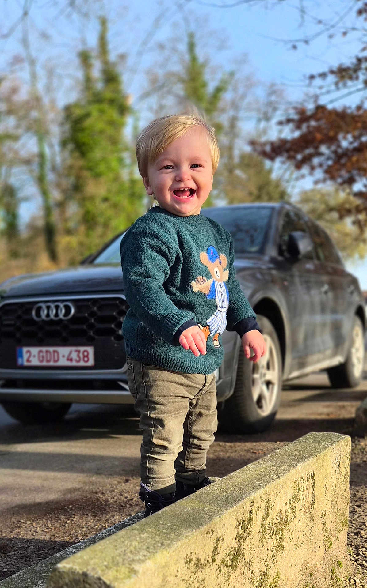 James participe au concours pour gagner de l'argent avec cette photo : toddler, child, smiling, sweater, bear_design, pants, boots, standing, concrete_ledge, car, audi, license_plate, outdoor, trees, daylight, happy, person, nature, road, blue_sky