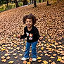 Autumn is registered to the contest to win money with this photo: autumn, black_sweater, blue_pants, casual_clothing, child, curly_hair, daylight, fall_leaves, happy, leafy_ground, nature, outdoor, park, person, playful, seasonal, smiling, tree, white_shoes, young