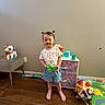 child, toddler, girl, smiling, standing, indoors, playroom, toy, unicorn, stuffed_animal, colorful, play_mat, dresser, chair, wood_floor, wall, socks, shorts, t_shirt, portrait
