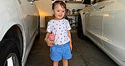 Gianna joined the competition — help win amazing prizes! child, toddler, girl, smiling, standing, portrait, garage, cars, vehicles, toy, pink_toy, pigtails, hair_bows, t_shirt, shorts, sneakers, indoor, family_photo, shelves, automotive