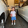 child, toddler, girl, smiling, standing, portrait, garage, cars, vehicles, toy, pink_toy, pigtails, hair_bows, t_shirt, shorts, sneakers, indoor, family_photo, shelves, automotive