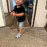 black_shirt, carpet, chair, child, curly_hair, cute, denim_shorts, doorway, floor, furniture, happy, home, indoor, living_room, person, shoes, smiling, toddler, walking, wood_floor
