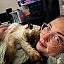 cat, person, glasses, smile, face, freckles, nose_ring, hand, blanket, indoor, furniture, technology, computer, screen, cuddling, pet, resting, cozy, comfort, closeup
