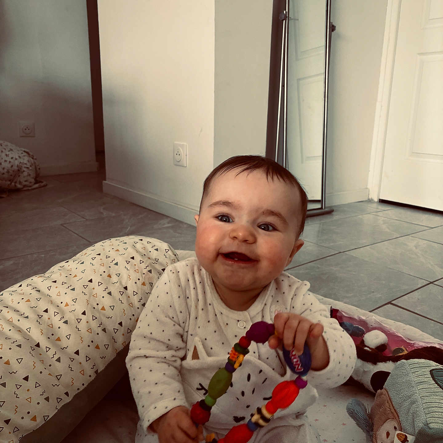 Michaëla a rejoint le concours — aidez-le/la à gagner de superbes lots ! baby, child, smiling, toy, playmat, pillow, plush_toy, onesie, indoor, floor, happy, cute, infant, person, home, childhood, colorful, sitting, face, baby_toys
