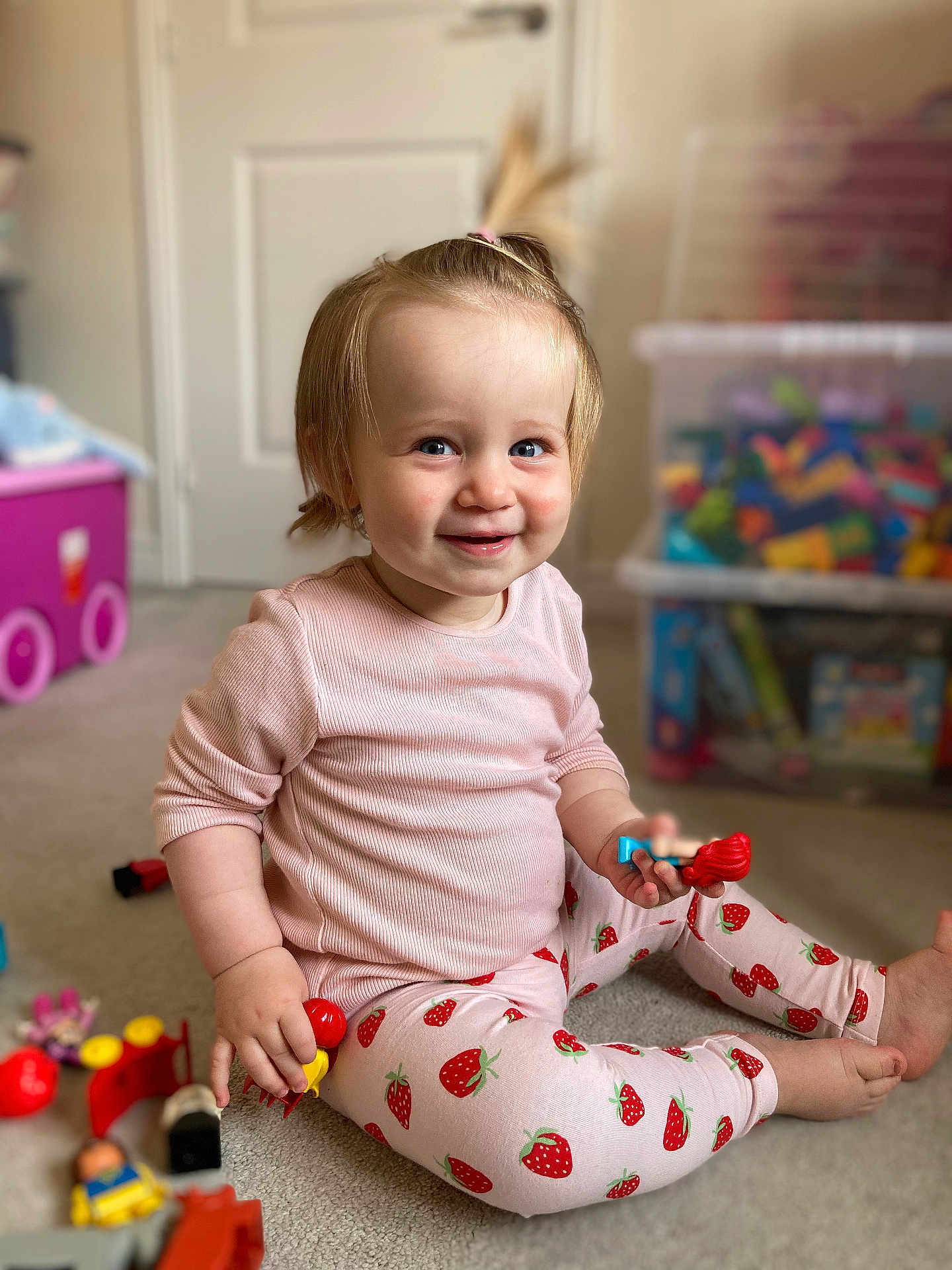 Aurora joined the competition — help win amazing prizes! toddler, child, smiling, indoor, toy, playtime, pink_clothing, strawberry_pattern, carpet, person, cute, happy, sitting, hair_tie, playroom, plastic_toys, floor, blue_eyes, small_hands, casual_wear