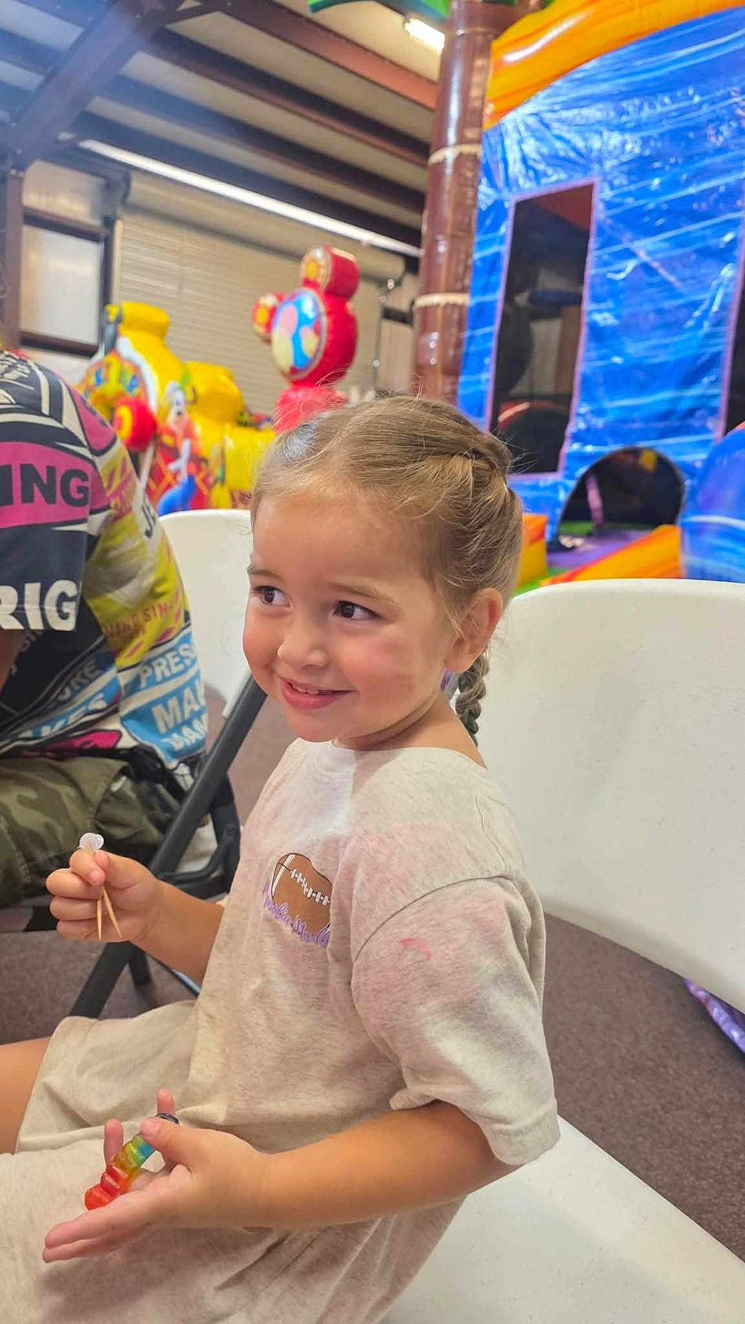 Baylynn is registered to the contest to win money with this photo: child, girl, smile, braided_hair, white_chair, gummy_candy, toothpicks, indoor, play_area, inflatable, colorful, fun, casual_clothing, happy, person, seat, background, recreation, event, cute