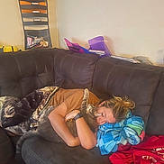 Simba is registered to the contest to win money with this photo: person, cat, couch, blanket, hugging, indoor, casual, relaxing, home, comfort, sleeping, affection, furniture, brown_clothing, watch, pillow, shelf, folder, red_clothing, multicolored_blanket