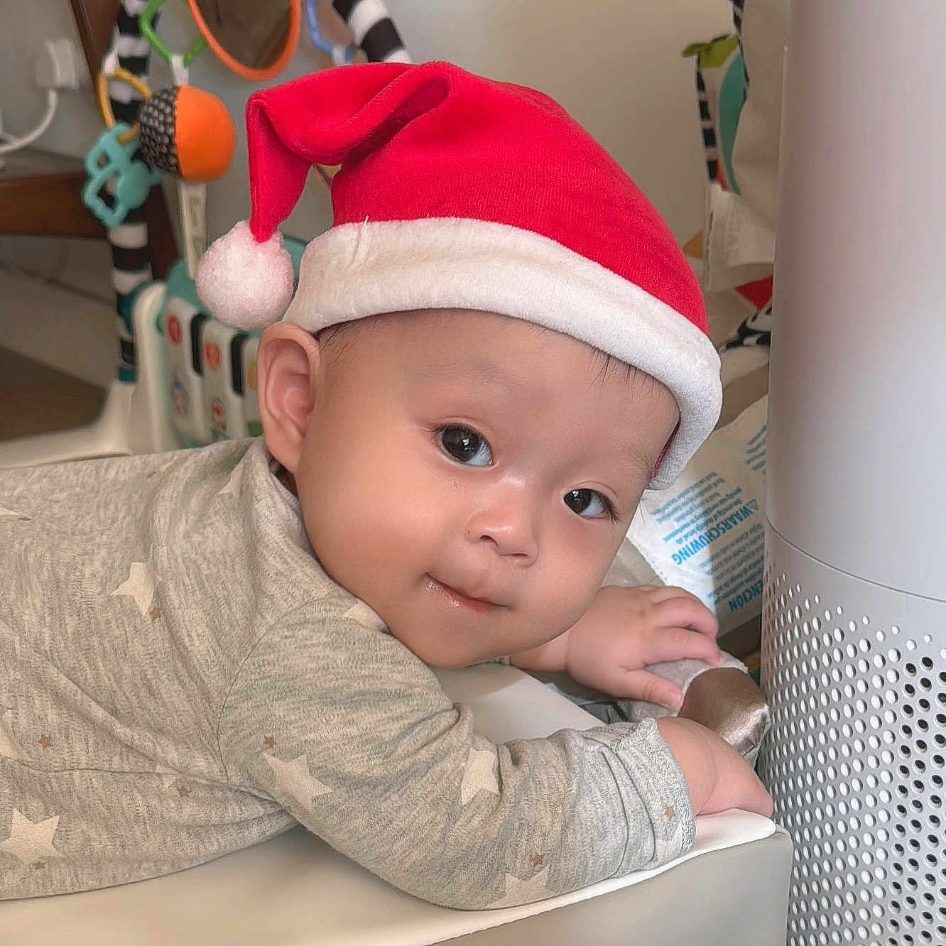 Alexander joined the competition — help win amazing prizes! baby, infant, santa_hat, gray_clothing, star_pattern, indoor, toy, air_purifier, curious_expression, face, child, cute, holiday, festive, soft_surface, hand, lying_down, headwear, smile, warm_lighting