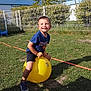 blue_sky, boy, child, fence, fun, grass, greenery, happy, hopper_ball, outdoor, play, recreation, shadow, shoes, shorts, smiling, sunlight, tree, tshirt, yellow_ball