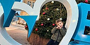 Yanis a rejoint le concours — aidez-le/la à gagner de superbes lots ! boy, building, child, christmas_tree, daylight, decorations, festive, fun, heart_shape, holiday, ornaments, outdoor, pavement, playful, pose, public_space, sculpture, sky, smiling, winter_clothing
