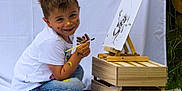Yanis a rejoint le concours — aidez-le/la à gagner de superbes lots ! art_supplies, boy, canvas, child, colorful, creative, easel, face_paint, grass, happy, jeans, nature, outdoor, paint, painting, playful, sitting, smiling, white_shirt, wooden_crate
