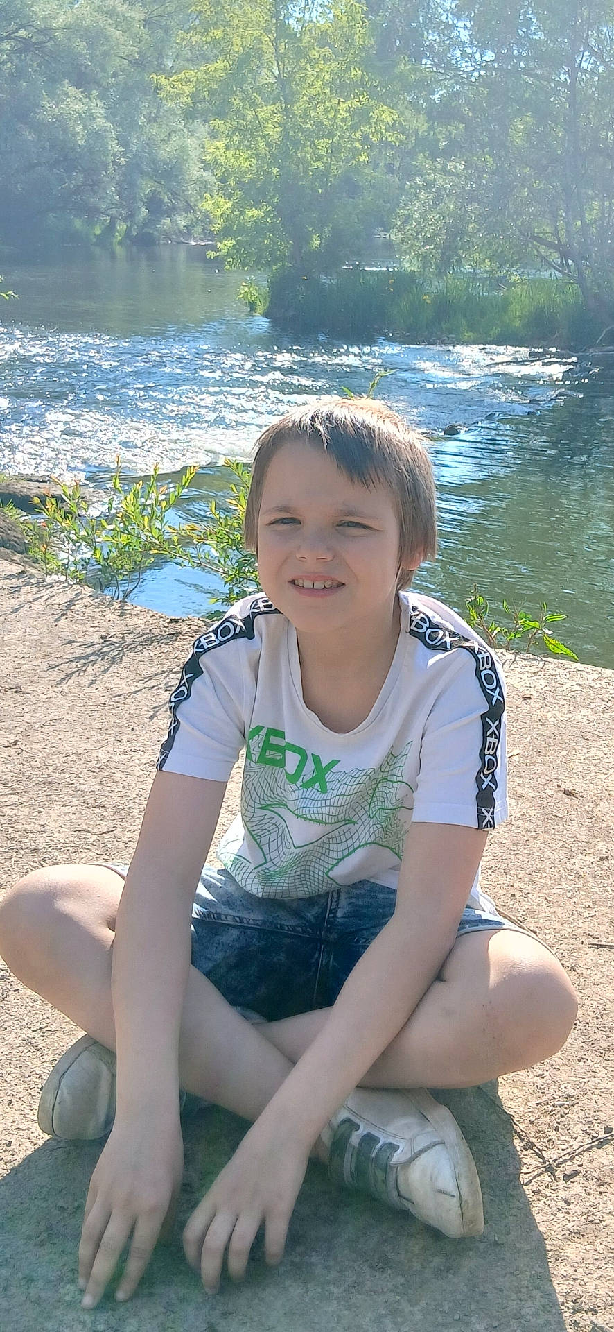 Cameronn participe au concours pour gagner de l'argent avec cette photo : boy, casual_clothing, child, daylight, greenery, nature, outdoor, park, portrait, relaxation, river, shoes, shorts, sitting, smile, sunlight, trees, tshirt, water, young