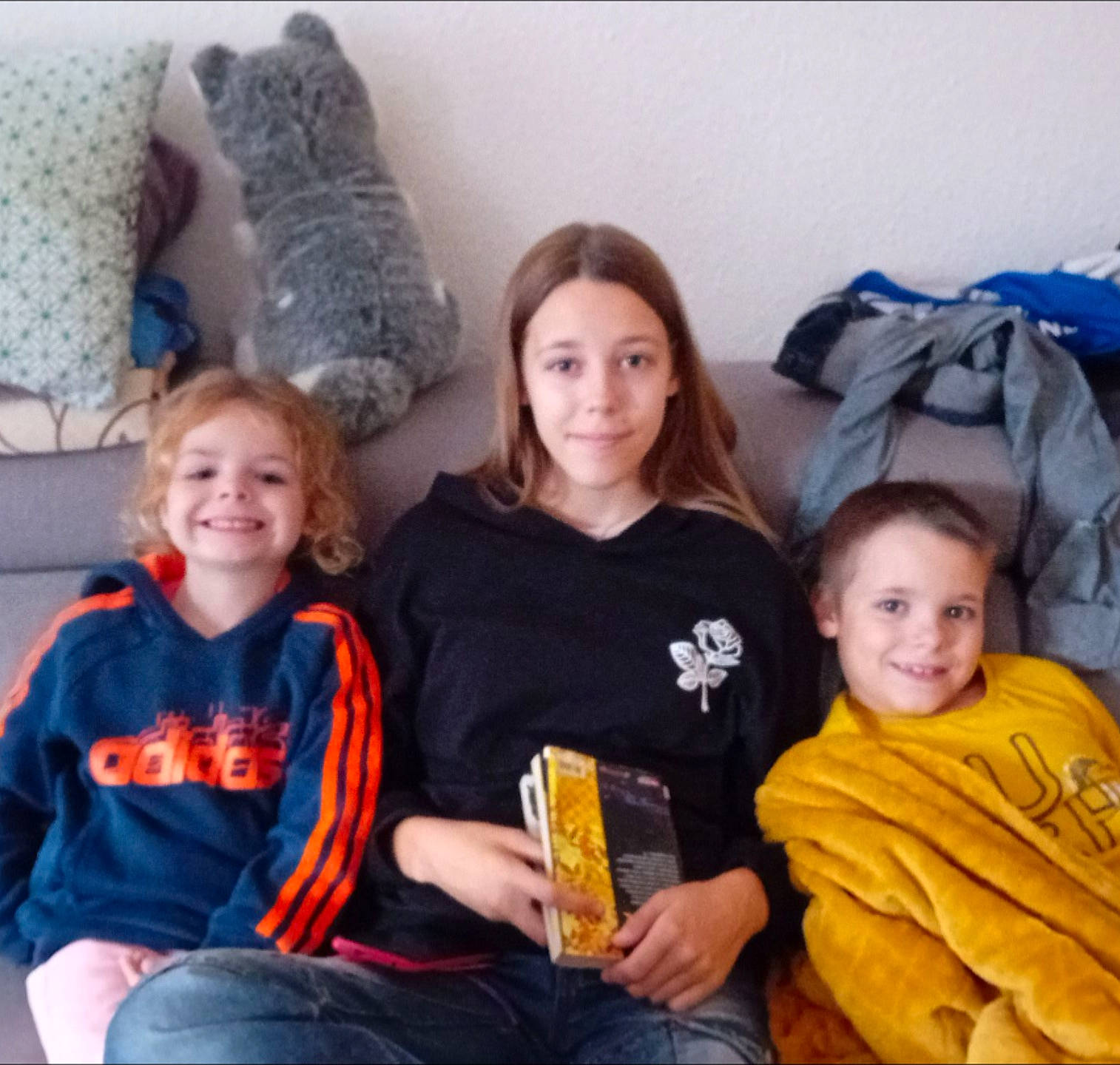 Cameronn participe au concours pour gagner de l'argent avec cette photo : blanket, book, casual_clothing, child, children, couch, cozy, family, hair, happy, indoor, living_room, pillow, portrait, relaxed, sitting, smile, stuffed_animal, three_people, young