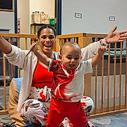 Ezeriel joined the competition — help win amazing prizes! toddler, woman, smiling, indoors, play_area, baby_gate, floor, carpet, clothing, polka_dot, red_pants, white_shirt, supporting, joyful, person, child, furniture, doorway, sitting, happy