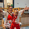 toddler, woman, smiling, indoors, play_area, baby_gate, floor, carpet, clothing, polka_dot, red_pants, white_shirt, supporting, joyful, person, child, furniture, doorway, sitting, happy